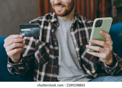 Cropped Young Man He Wear Brown Shirt Use Mobile Cell Phone Hold Credit Bank Card Shopping Online Order Delivery Booking Tour Sitting On Blue Sofa In Own Living Room Apartment Stay Home Indoor Flat On