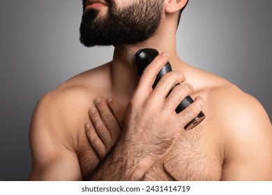 Cropped of young muscular man trimming his thick beard or shaving neck with electric shaver, isolated on grey studio background. Beard care tools concept