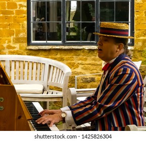 Cropredy, Oxfordshire. England - June 15, 2019: Pony Tailed Man In Straw Boater And Striped Jacket, Entertains The Crowd, By Playing A Piano, Outside At The Annual Village Fete.