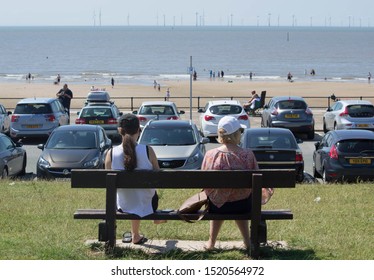 Crosby, Merseyside/UK - July 23rd 2019: day at the beach, women sat on bench overlooking a car park, sandy beach and distant sea with wind turbines and holidaymakers enjoying the hot weather 