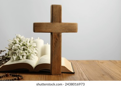 Cross, Bible, rosary beads, flowers and church candles on wooden table against light background. Space for text
