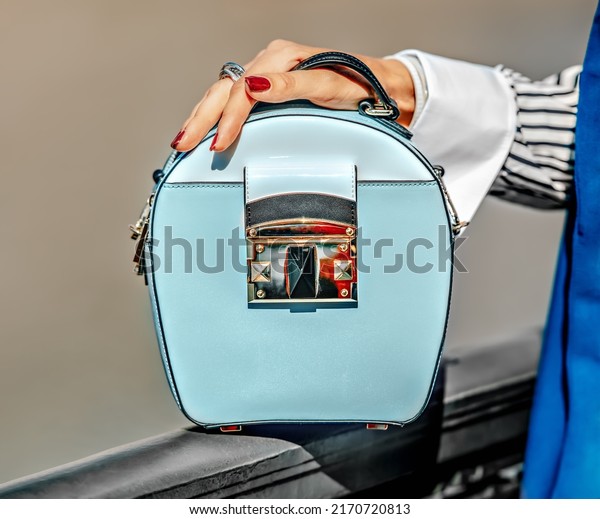 Cross body bag with\
handle. Leather handbag of light blue color with a gold lock.\
Stylish accessories for a business woman. The woman is holding a\
small bag in her hands.
