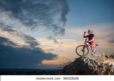 Cross Country Biker Drinking Water On Top Of A Mountain With Bike In The Evening, Sky Background. Side View