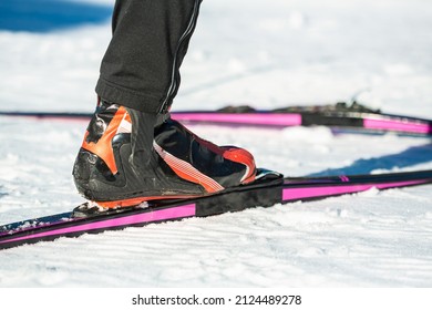 Cross country skier legs standing on skis, putting the toe of the ski boot in the notch on the ski, and hooking the metal bar on the boot.