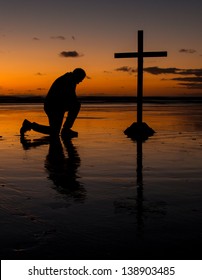 Cross with a man kneel in prayer on a beach at sunset.