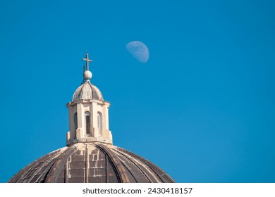 A cross on a top of catholic church dome and the Moon on background of blue sky