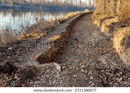 cross section road built or reconstructed, probably cycle path, footpath along river. transition from an unpaved or gravel surface to freshly laid concrete base. bordered side by concrete curbs. finished asphalt section continues.  surroundings are an autumn or winter landscape with fallen leaves and dry vegetation