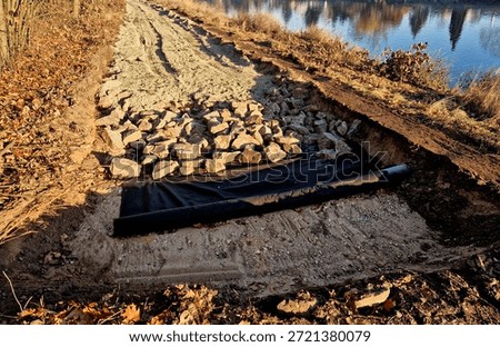 cross section road built or reconstructed, probably cycle path, footpath along river. transition from an unpaved or gravel surface to freshly laid concrete base. bordered side by concrete curbs. finished asphalt section continues.  surroundings are an autumn or winter landscape with fallen leaves and dry vegetation
