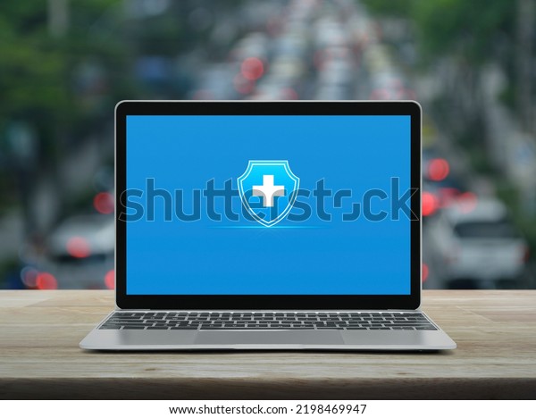 Cross shape with shield flat icon on modern\
laptop computer screen on wooden table over blur of rush hour with\
cars and road in city, Business healthy and medical care insurance\
online concept