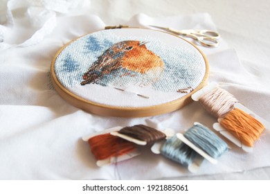 cross stitch process in the hoop multicolored bird, white canvas, floss threads on bobbins, needle, needlework and cross stitch concept