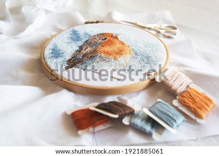 cross stitch process in the hoop multicolored bird, white canvas, floss threads on bobbins, needle, needlework and cross stitch concept 