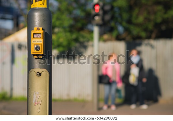 cross walk remote and its sign push button on\
pole with red stop red light and two girls waiting in background,\
pedestrians