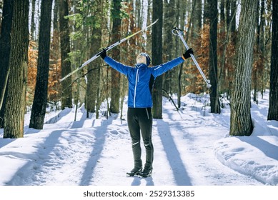 Cross-country skier in a blue jacket raises their arms in triumph, enjoying a winter day in a snowy forest. - Powered by Shutterstock