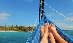 Crossed Feet Of A Man And Woman Wearing Silver Toe Ring Relaxing In A Blue Spotted Hammock Hanging From The Rigging Of A Sailboat Floating Over Aquamarine Water Facing A Lush, Palm Tree Covered Beach.