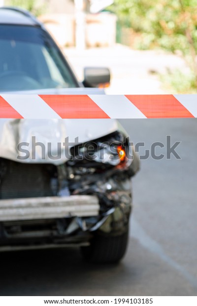 Crossed red white warning tape in front of car\
accident. Broken hood of car on road fenced by Red warning tape.\
Broken Bumper and Car Headlights with Light On road surface asphalt\
with nobody