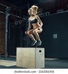 Crossfit young beautiful woman jumping onto a box as part of exercise routine. Fitness woman doing box jump workout at gym. Box jumping workout at modern gym. Powerful attractive muscular woman.