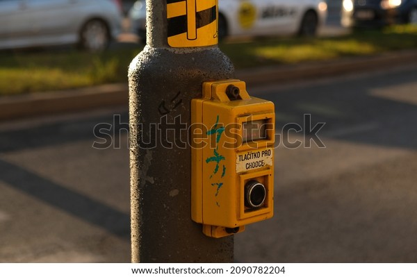 Crosswalk button for pedestrian with light warning.\
Defocused background with Cars drive along road and bokeh from\
headlights. The inscription in Czech -Tlačítko pro chodce- Button\
for pedestrians.  