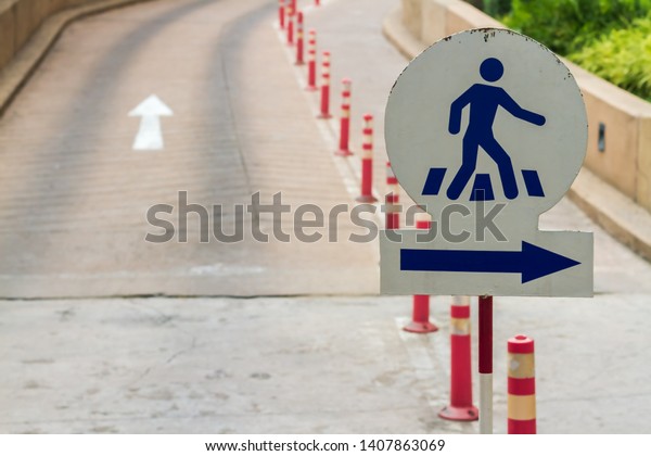 Crosswalk symbol and blue arrow sign shown on white\
metal plate with group of orange traffic pillars and old concrete\
road background. 