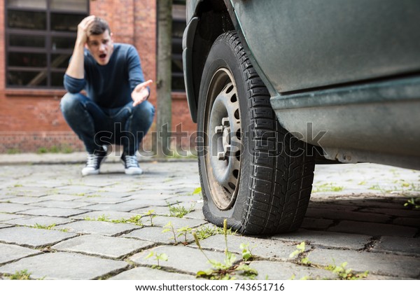 Crouched Worried Young Man With Hand On Head\
Pointing At Punctured Car\
Tire