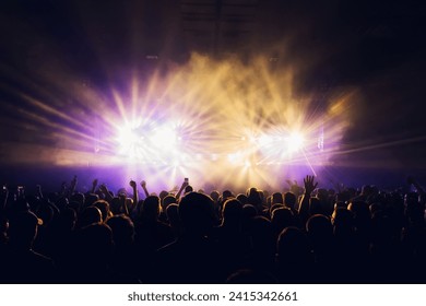 Crowd dancing and having fun at night on a music festival. There are stage purple and yellow lights. Silhouettes of people having fun, dancing on music festival. Nightlife. - Powered by Shutterstock