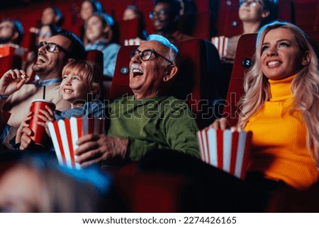 A crowd of happy spectators are in the movie, sitting in the chairs laughing at the movie they are watching and enjoying popcorn and drinks.