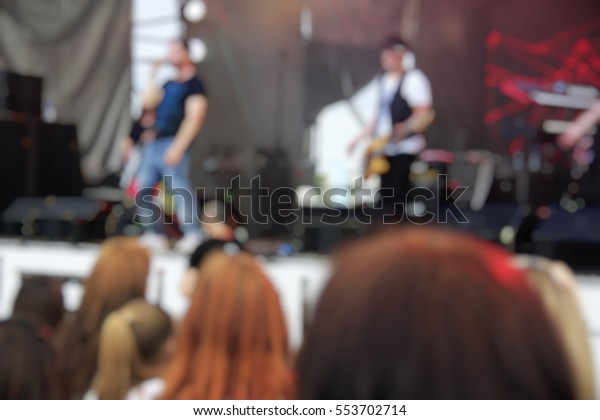 A crowd of people at a concert in the park.\
Near the scene. Performance of the musical group. Artists. Blurred\
background