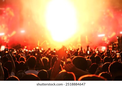 Crowd of people enjoys and cheering music concert in front of bright stage lights. Happy audience dance and raise hands.