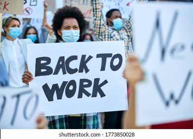 Crowd of people protesting against job loss due to COVID-19 pandemic. Focus is on African American woman holding a placard with back to work inscription. 