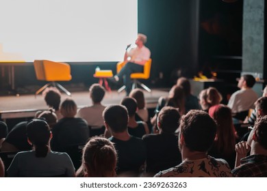 Crowd sitting in hall against blurred stage, while speaker showing presentation on big screen and speaking into mic during seminar - Powered by Shutterstock