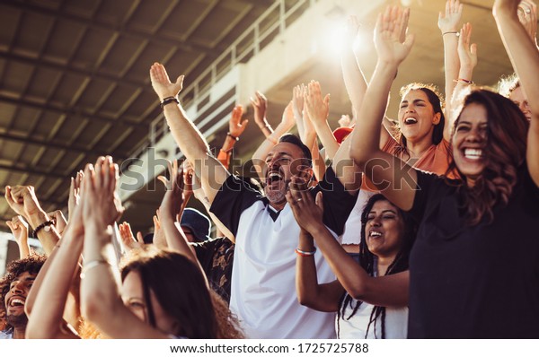 Crowd of sports fans cheering during a match\
in stadium. Excited people standing with their arms raised,\
clapping and yelling to encourage their\
team.