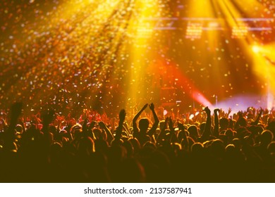 A Crowded Concert Hall With Scene Stage Orange And Yellow Lights, Rock Show Performance, With People Silhouette, Colourful Confetti Explosion Fired On Dance Floor Air During A Concert Festival
