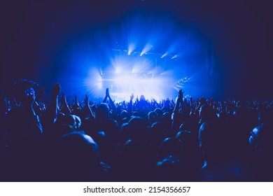 A crowded concert hall with scene stage lights in blue tones, rock show performance, with people silhouette, on a dance floor air during a concert festival - Shutterstock ID 2154356657