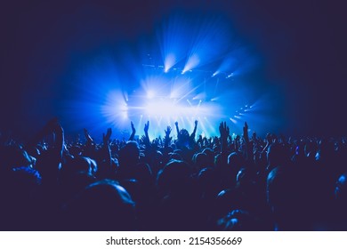 A crowded concert hall with scene stage lights in blue tones, rock show performance, with people silhouette, on a dance floor air during a concert festival - Shutterstock ID 2154356669