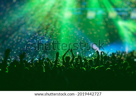 A crowded concert hall with scene stage green lights, rock show performance, with people silhouette, colourful confetti explosion fired on dance floor air during a concert festival