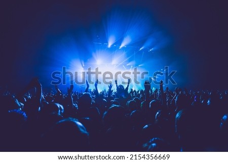 A crowded concert hall with scene stage lights in blue tones, rock show performance, with people silhouette, on a dance floor air during a concert festival