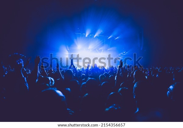 A crowded concert hall with\
scene stage lights in blue tones, rock show performance, with\
people silhouette, on a dance floor air during a concert\
festival