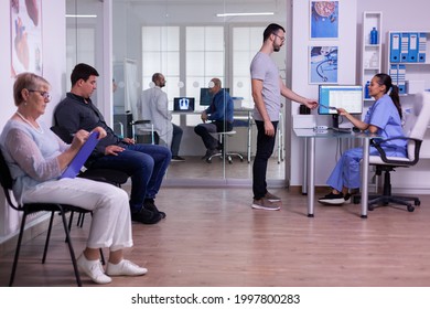 Crowded Hospital Waiting Area With People Filling Form For Medical Consultation, Young Man Giving Xray To Nurse, Making New Appointment. Specialist Doctor Consulting Senior Man In Examination Room.