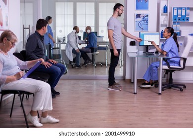 Crowded Hospital Waiting Area With People Filling Form For Medical Consultation, Young Man Giving Xray To Nurse, Making New Appointment. Specialist Doctor Consulting Senior Man In Examination Room.