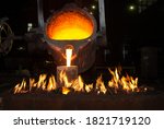 Crucible pouring molten iron into a vessel heated by flames in a foundry.