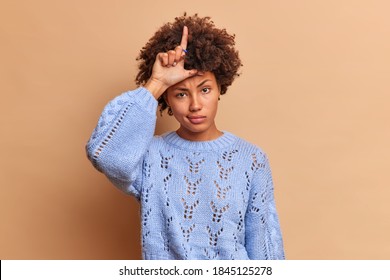 Cruel Rude Serious Afro American Woman Makes Lose Gesture Keeps Finger Raised Over Forehead Insults Someone Makes Fun Of People Wears Blue Sweater Isolated Over Beige Background. Body Language