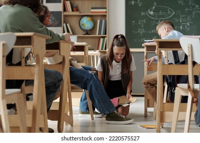 Cruel schoolkids laughing at their classmate standing on knees in aisle between desks and picking her copybooks off the floor