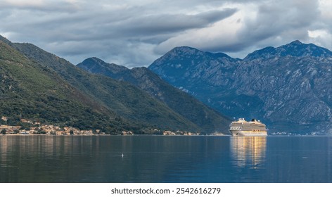 A cruise ship with illuminated lights sails into Kotor Bay in the early morning under a moody sky. Majestic mountains surround the calm waters, creating a serene and scenic atmosphere. - Powered by Shutterstock