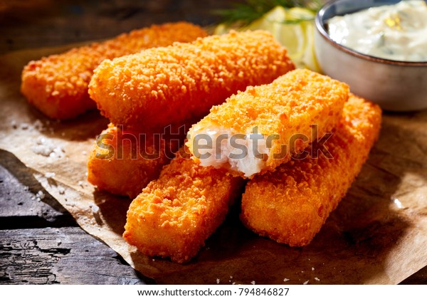 Crumbed golden fried fish fingers in sticks\
served on brown paper on a rustic wood counter with tartare sauce\
in a close up view with one broken\
open