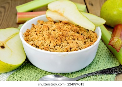 Crumble with pears and rhubarb in a white bowl on a napkin, spoon on a wooden boards background