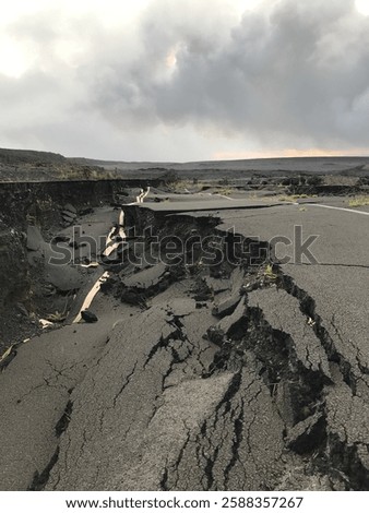 A crumbled road split by an earthquake, jagged asphalt lifted and fractured. Deep cracks snake across the surface, with chunks of pavement scattered on the dusty ground.