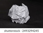 A crumpled white paper ball against a dark background. The image highlights the textures and folds of the paper, symbolizing waste, mistakes, or recycling.