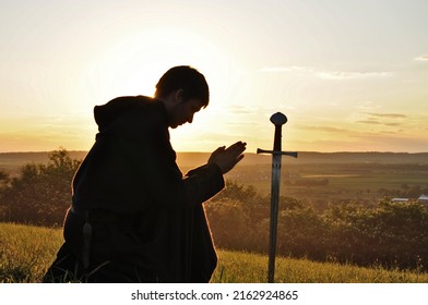 Crusader Knight Praying And Kneeling Down To A Sword Like A Cross. Religious Scene In Medieval Europe. Excalibur And King Arthur Saga.