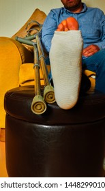 Crutches Along Side A Person Sat Down, With There Foot Bandaged Resting On A Foot Stool At Home.