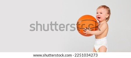 Crying baby with big basketball ball. Little cute toddler girl, child in diaper posing isolated on grey studio background. Concept of family, childhood, motherhood, life, birth. Banner