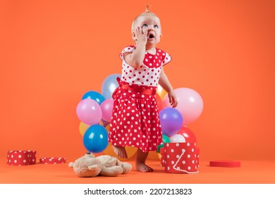 Crying Baby Girl In Red Dress Upset With Birthday Gifts. Studio Photo On Orange Background